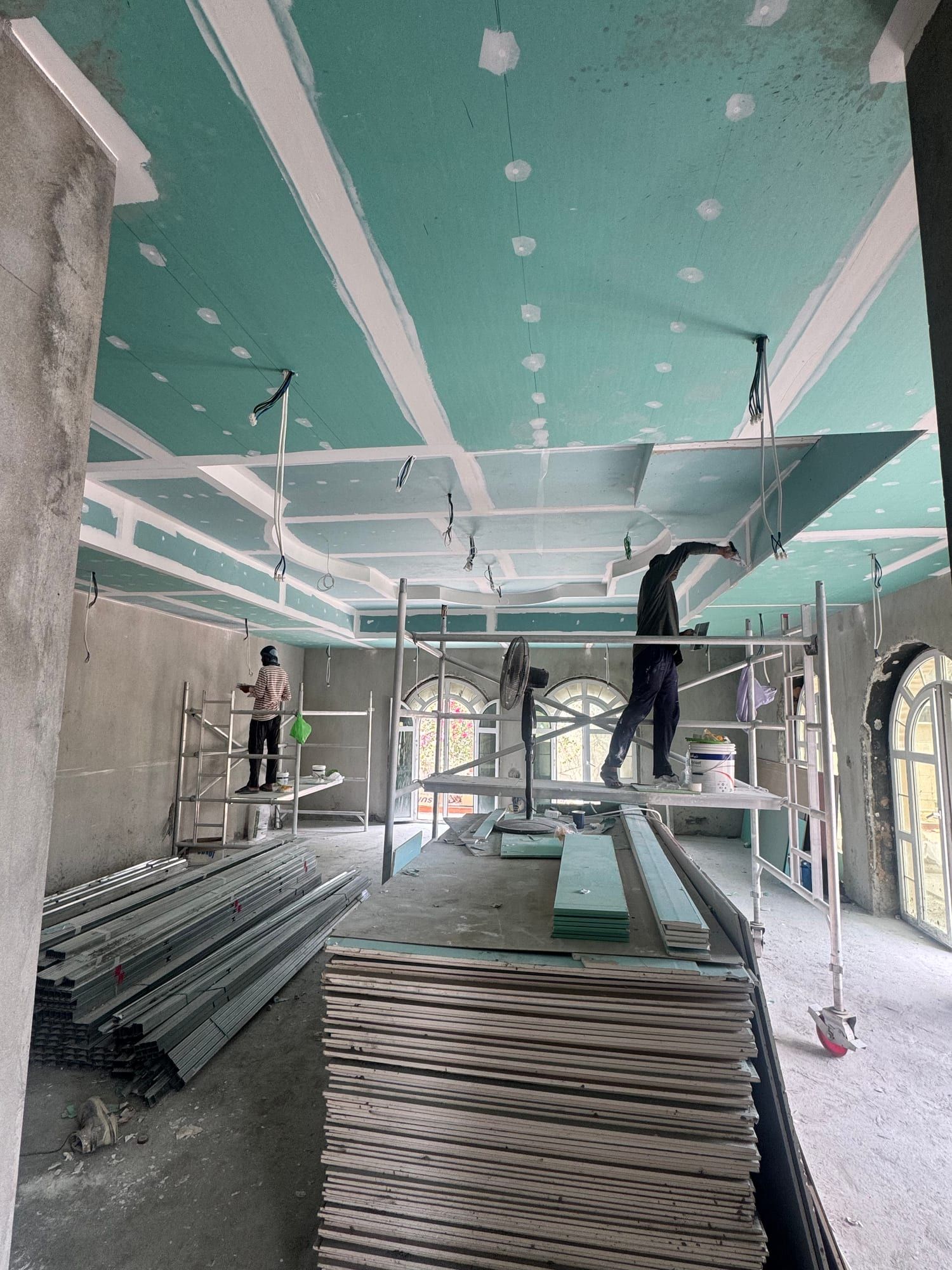 Workers on mobile scaffolds fixing green boards to a stepped ceiling, with stacks of boards on the floor
